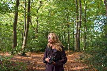 A woman dressed in natural colors walks through a leafy autumn fall forest whilst holding a dslr camera. She looks relaxed and natural as she walks through the beautiful autumnal woodland.