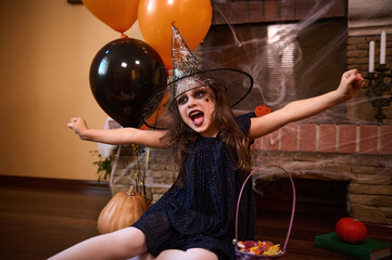 Mischievous child girl in wizard's hat, little enchantress, casts spells sitting on the floor, surrounded by a cobweb-covered fireplace, black and orange air balloons, pumpkins and Halloween treat