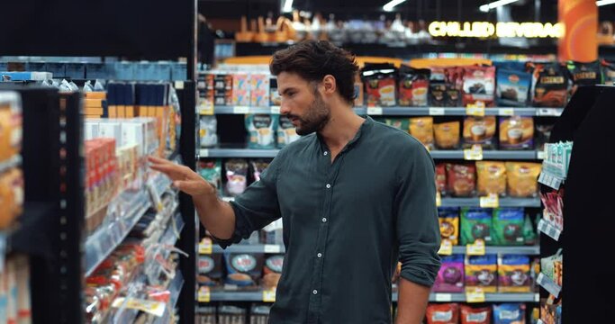 Young dark-haired bearded multi-ethnic sporty guy in casual clothes buys goods, reads the composition on the package of cereals, while shopping for goods in the grocery department of the supermarket