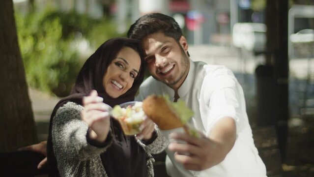 Young muslim couple showing their lunch to camera and eating it. Handsome bearded man and lovely woman in hijab enjoying their meal outside on summer day. Front view. Relationship, lifestyle concept.