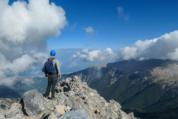 Rear view of man with climbing helmet on cloud covered mountain summit of Mytikas Mount Olympus, Mt Olympus National Park, Macedonia, Greece, Europe. View of rocky ridges and Mediterranean Aegean Sea