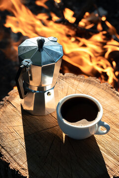 Coffee outdoors. Cup of black coffee and a moka pot on a stump in front of a campfire