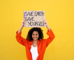 Cheerful African American young woman holding a sign that says: Save earth save yourself. Environmentalism, not planet b