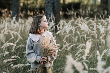 Portrait of thoughtful caucasian teen girl with closed eyes in profile wearing casual trendy clothes, holding bouquet of dry pampas grass in her hands and staying in autumn forest in golden hour.