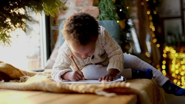 Merry Christmas. A little African American boy in a New Year's sweater lies by the window and writes a letter to Santa Claus