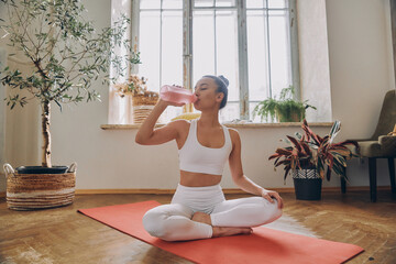 Beautiful young woman drinking water after training while sitting on the exercise mat at home