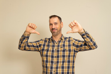 Studio portrait of funny proud man in his 40s pointing thumbs to himself, saying he's really cool, looking successful and confident, wearing shirt, standing over beige background, with copy space.