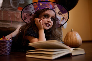 Inspired little girl dressed as a witch in a wizard's hat, cutely smiles looking mysteriously at camera while reading a sorcery spell book, lying on the floor against a spider web covered fireplace