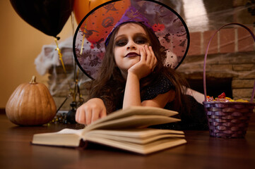 Adorable little child girl with face art make-up, in wizard hat, lying on the floor and reading...