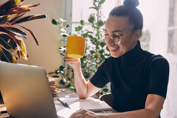 Happy woman using laptop and enjoying hot drink while sitting at her working place in office