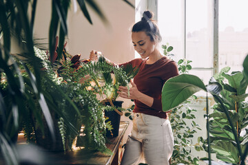 Happy young woman watering houseplants while standing at the domestic room - 538677071