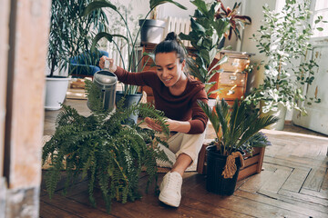 Confident young woman watering houseplants while sitting on the floor at home