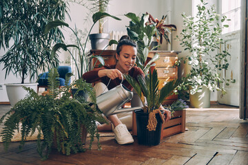 Beautiful young woman watering houseplants and smiling while sitting on the floor at home