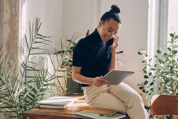 Happy woman talking on mobile phone and using digital tablet while sitting on the desk in office