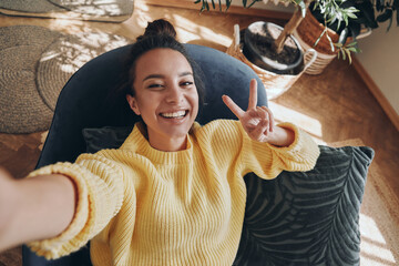 Top view of happy young woman making selfie and gesturing while relaxing in chair at home
