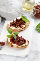 Sandwiches with cottage cheese, ricotta cheese, dried tomatoes in oil and basil. Bruschetta, snack, appetizers. Toasts on white board and marble background.