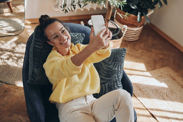 Happy young woman making selfie by her smart phone while relaxing in a chair at home