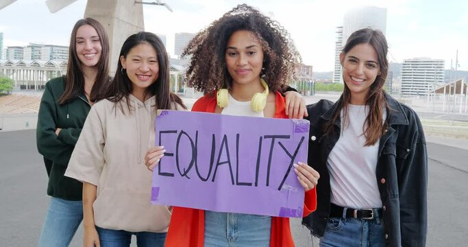 Group of cheerful multiethnic girls looking at camera holding a sign that says: Equality. Feminism, empowered woman
