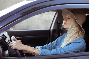 Fototapeta na wymiar Young woman with long fair hair in denim shirt, beige floppy hat, sitting in black car, holding steering wheel, driving.