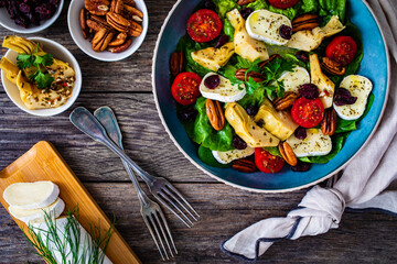 Fresh vegetable salad with cheese - camembert, artichoke, lettuce, cherry tomatoes, cucumber,  pekan nuts, arugula and  spinach  on wooden table
