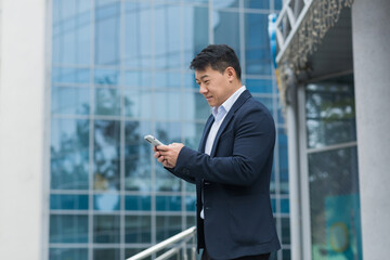 Successful Asian businessman in business suit walking outside near office building, male manager director using phone, smiling and happy reading online message.
