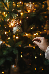 Happy New Year! Burning sparkler in female hand on background of christmas tree lights in dark...