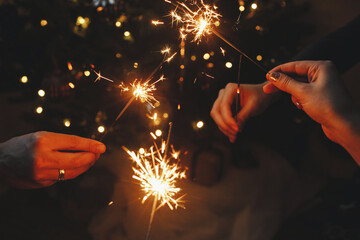 Happy New Year! Hands holding fireworks against christmas lights in dark room. Atmospheric holiday. Friends celebrating with burning sparklers in hands on background of stylish illuminated tree