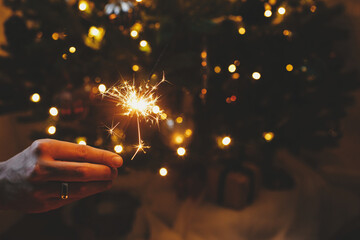 Happy New Year! Burning sparkler in male hand on background of christmas tree lights in dark room....