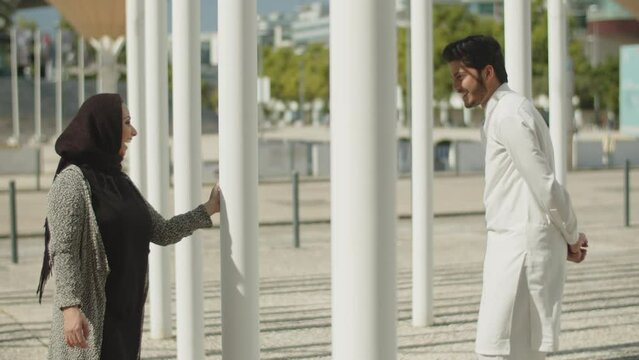 Young muslim couple playing hide-and-seek game between columns on sunny day. Arabic man in traditional clothes and woman in hijab having fun, dancing and flirting outside. Romance, ethnicity concept. 