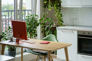 Cozy workplace for freelancer with stylish red monitor on table near with large window and kitchen stove. Modern home office designed to work comfortably for copywriter with lots of houseplants