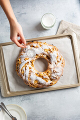 A woman's hand sprinkles powdered sugar on a yeast braided ring bun with apple-cinnamon filling, lying on baking paper on a baking sheet on a concrete gray background. Top view.