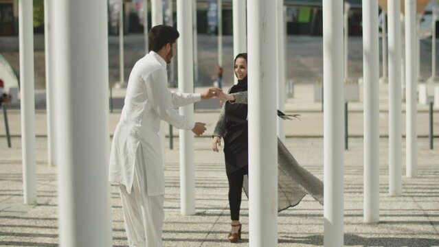 Happy young muslim couple dancing between columns on sunny day. Dark-haired man in white clothes and woman in hijab walking between columns, having fun together. Long shot. Romance, culture concept. 