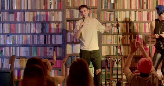 Stand-up comedian stage with books background. He runs out, hits microphone, greets audience. Broadcasting monologue comedy. Presenter microphone transmission. CZ, Prague, 8.10.22, Evropska