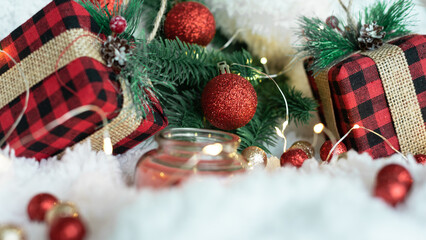 Decor from gifts in a checkered wrapper, branches of a Christmas tree, lights of a New Year's garland, a red candle, red and gold balls on a white fluffy background. Place for text. 