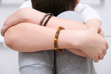A sad boy sitting on the floor, hiding his face and hugging knees, ethnic braselets on his hands