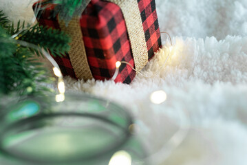 Focus on a gift in a plaid package decorated with a burlap ribbon, a warm-colored garland and a candle in a glass on a white soft background. Cozy holidays. New Year holidays 2023.