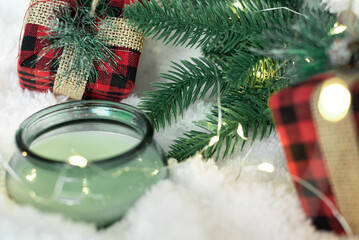Green candle,gifts in a checkered cloth wrapper, yellow lights on a garland, a sprig of a Christmas tree on a white soft background. Focus on the gift. Postcard for the New Year and Christmas.