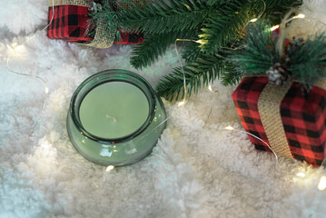 Green candle in glass and gifts in a checkered cloth wrapper, yellow lights on a garland, a sprig of a Christmas tree on a white soft background. Top view