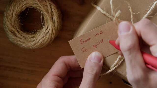 Man writing a gift tag with text FROM ME TO YOU on eco friendly wrapped gift box, close up view.