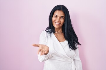 Mature hispanic woman standing over pink background smiling cheerful offering palm hand giving assistance and acceptance.
