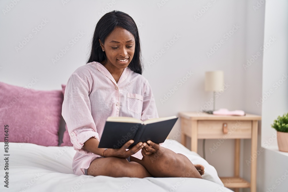 Wall mural young african american woman reading book sitting on bed at bedroom