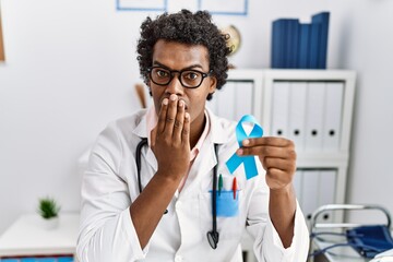 African doctor man holding blue ribbon covering mouth with hand, shocked and afraid for mistake....