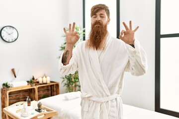 Fototapeta na wymiar Redhead man with long beard wearing bathrobe at wellness spa relax and smiling with eyes closed doing meditation gesture with fingers. yoga concept.