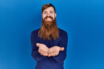 Redhead man with long beard wearing casual blue sweater over blue background smiling with hands palms together receiving or giving gesture. hold and protection