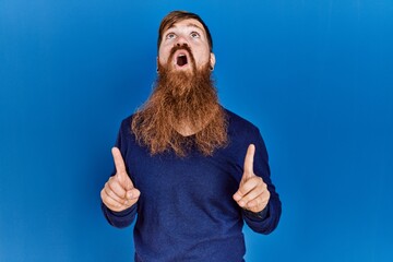 Redhead man with long beard wearing casual blue sweater over blue background amazed and surprised looking up and pointing with fingers and raised arms.