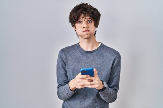 Young man using smartphone typing a message puffing cheeks with funny face. mouth inflated with air, catching air.