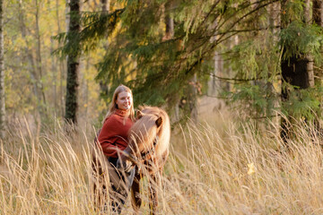 Naklejka na ściany i meble Young woman with Icelandic horse in tall grass during golden hour. Sun is setting and still visible Autumn scenery