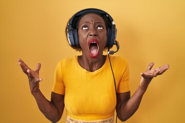 African woman with curly hair standing over yellow background wearing headphones crazy and mad...