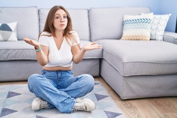 Fototapeta premium Young caucasian woman sitting on the floor at the living room clueless and confused expression with arms and hands raised. doubt concept.