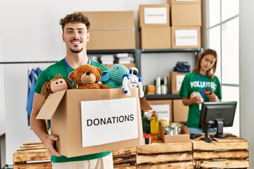 Young hispanic volunteer couple working at charity center. Man smiling happy holding box with toys...